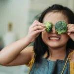 Asian chinese female child act cute with hand holding broccoli putting in front of her eyes with smiling face at kitchen